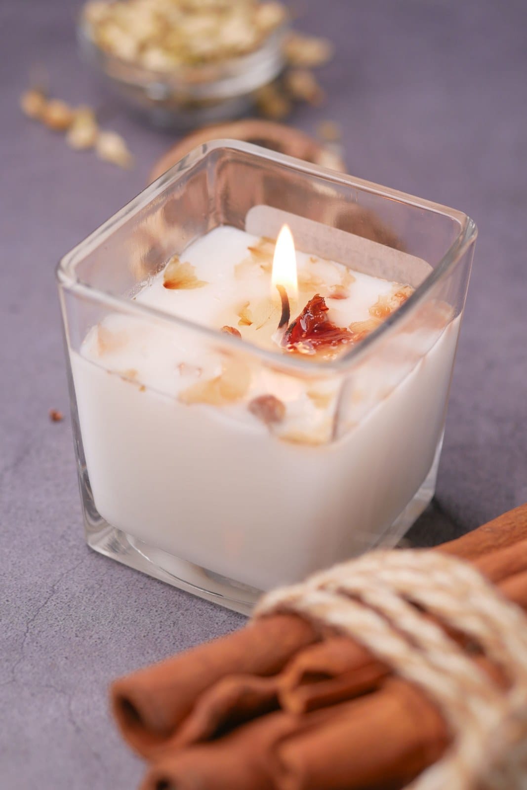 Lit candle with dried petals in a square glass jar beside cinnamon sticks tied with twine