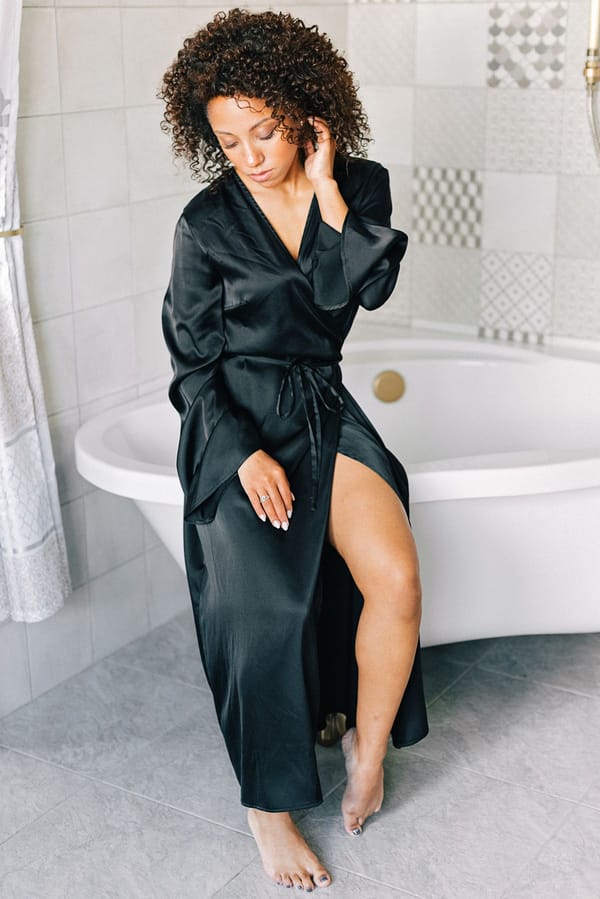 Woman in black silk robe sitting on a bathtub edge in a tiled bathroom