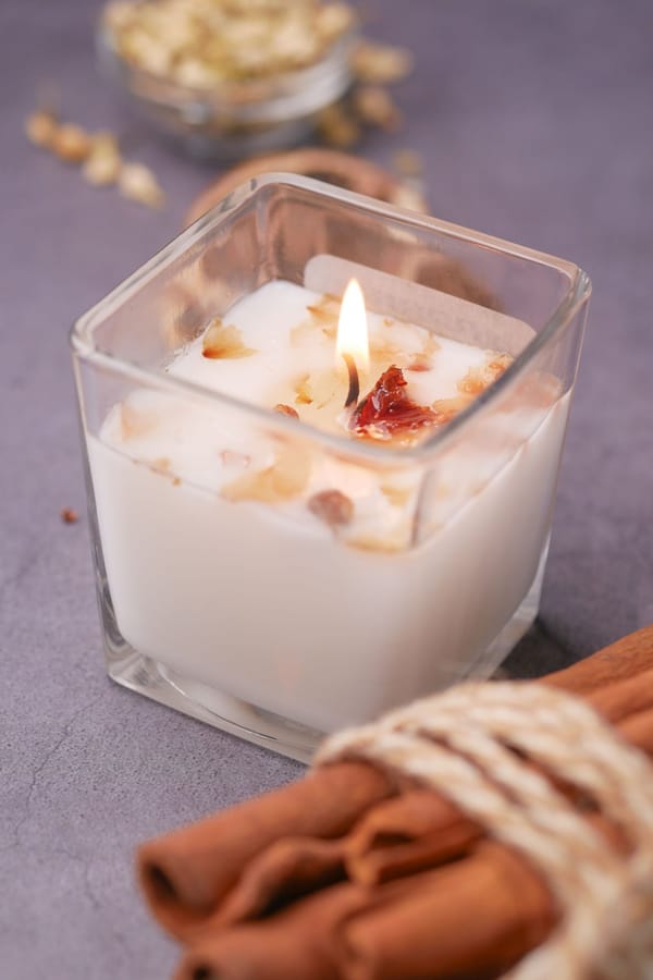 Lit candle with dried petals in a square glass jar beside cinnamon sticks tied with twine