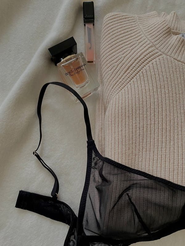 Sheer black designer bra laid flat beside a cream knit and perfume